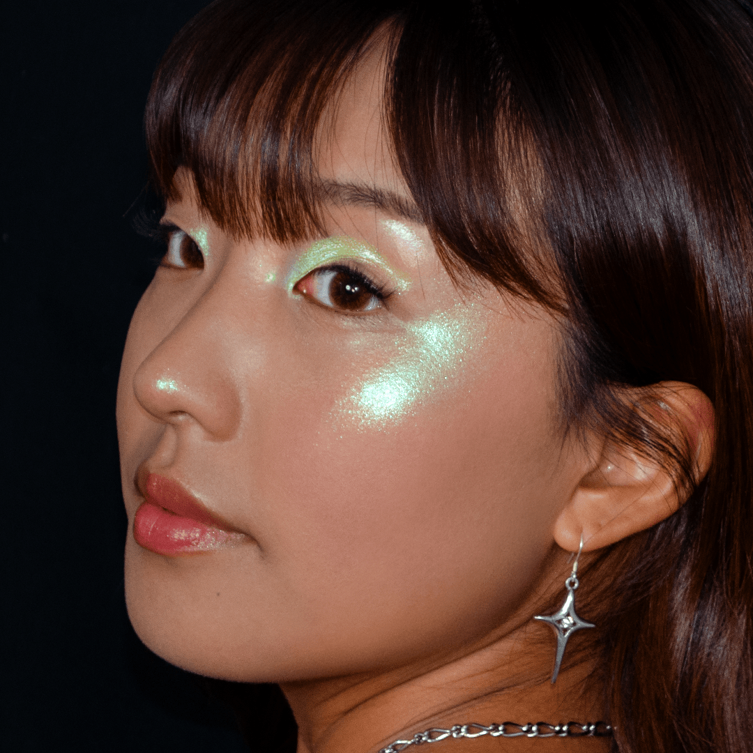 A woman with green eyeshadow and shiny  green cheeks wears star earrings and looks over her shoulder in a dark setting.