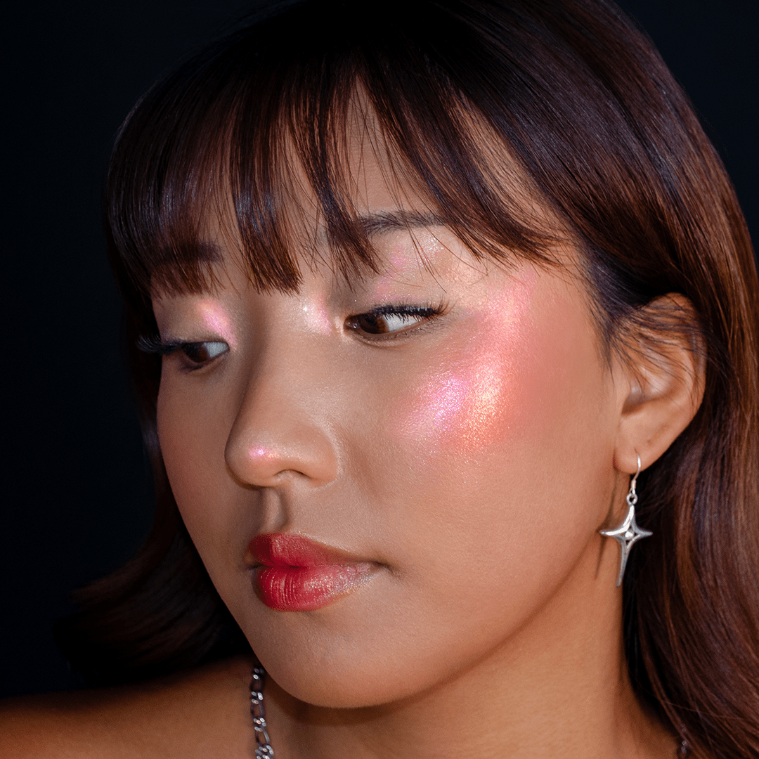 A woman with shimmery pink makeup on her cheeks and eyes wears a star earring and looks downward.