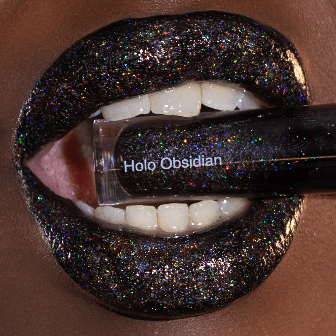 Close-up of lips with black glitter lipgloss holding a tube labeled Holo Obsidian with teeth.