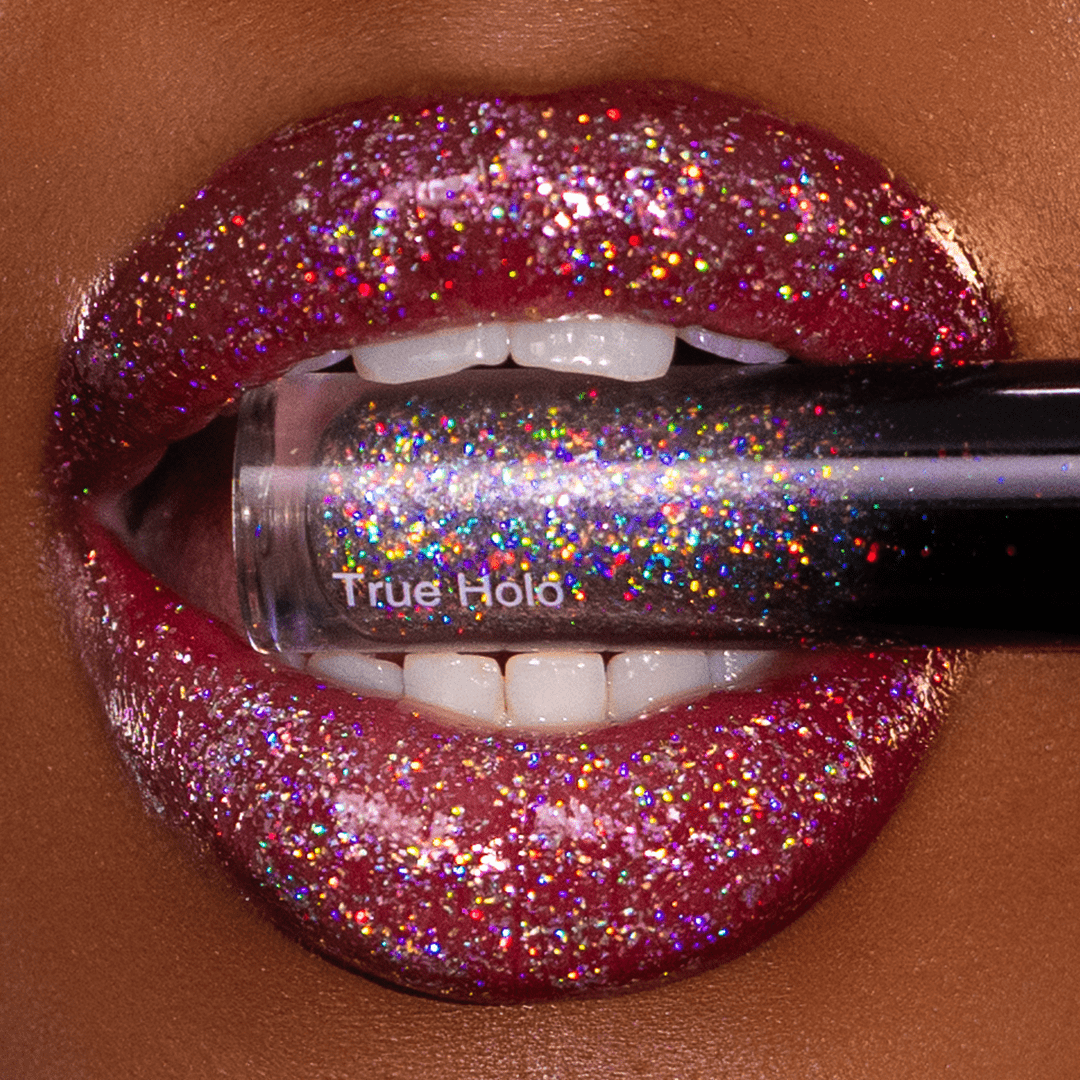 Glittery lips hold a prismatic tube of True Holo between white teeth.