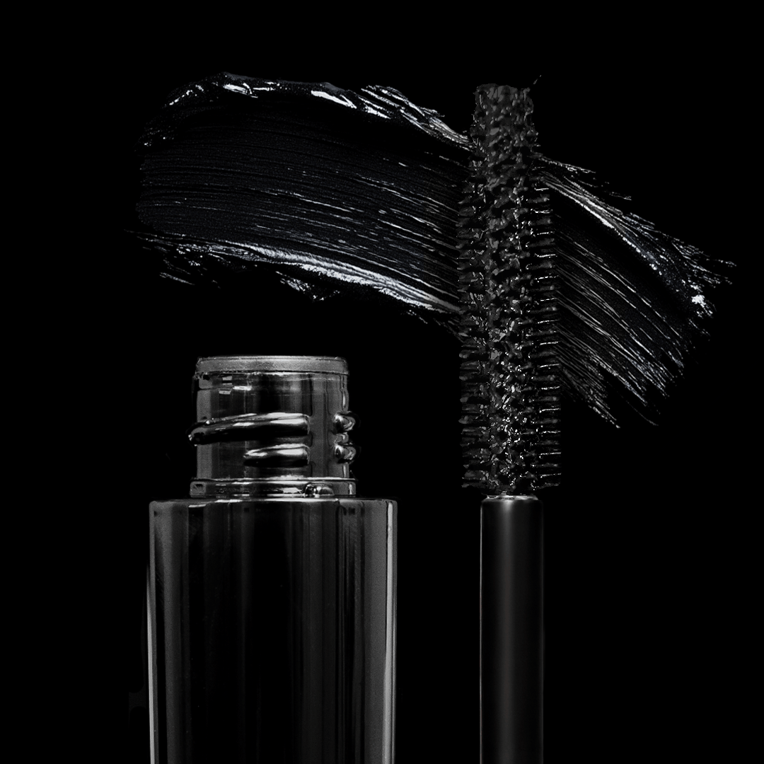 A black mascara tube and open wand with a glossy swipe of black sit on a solid black background.