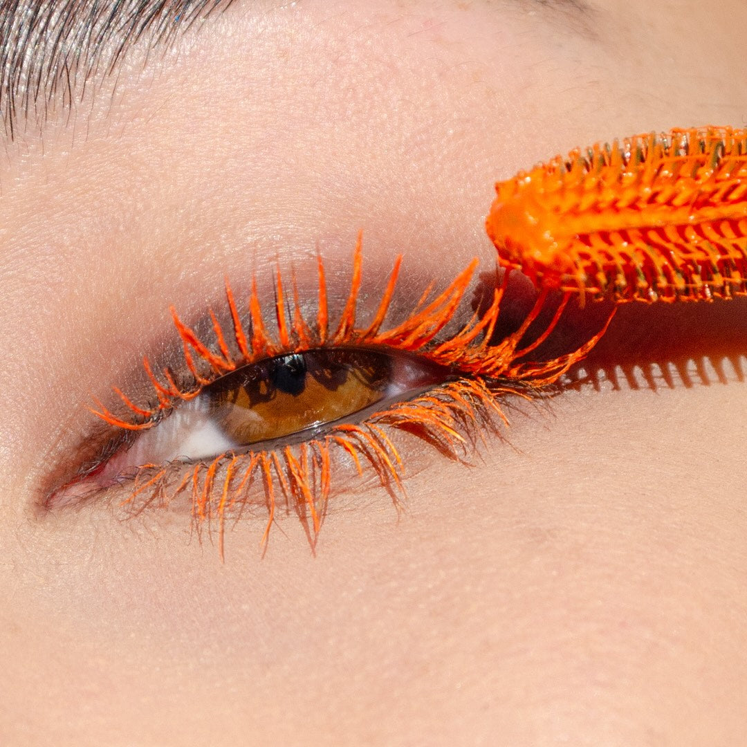 A close-up of an eye with bright orange mascara being applied to the upper lashes.