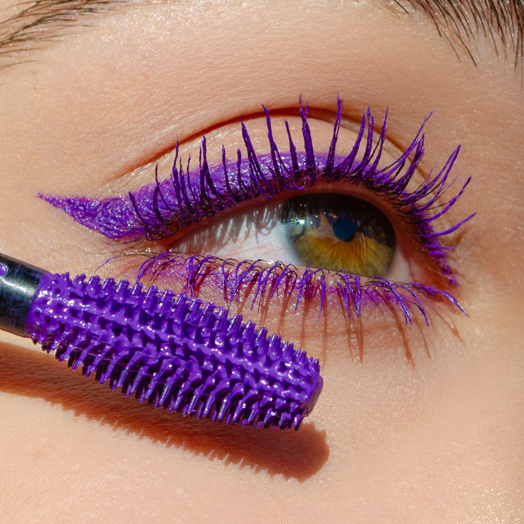 Close-up of an eye with purple mascara on volumized lashes and a purple mascara wand visible.