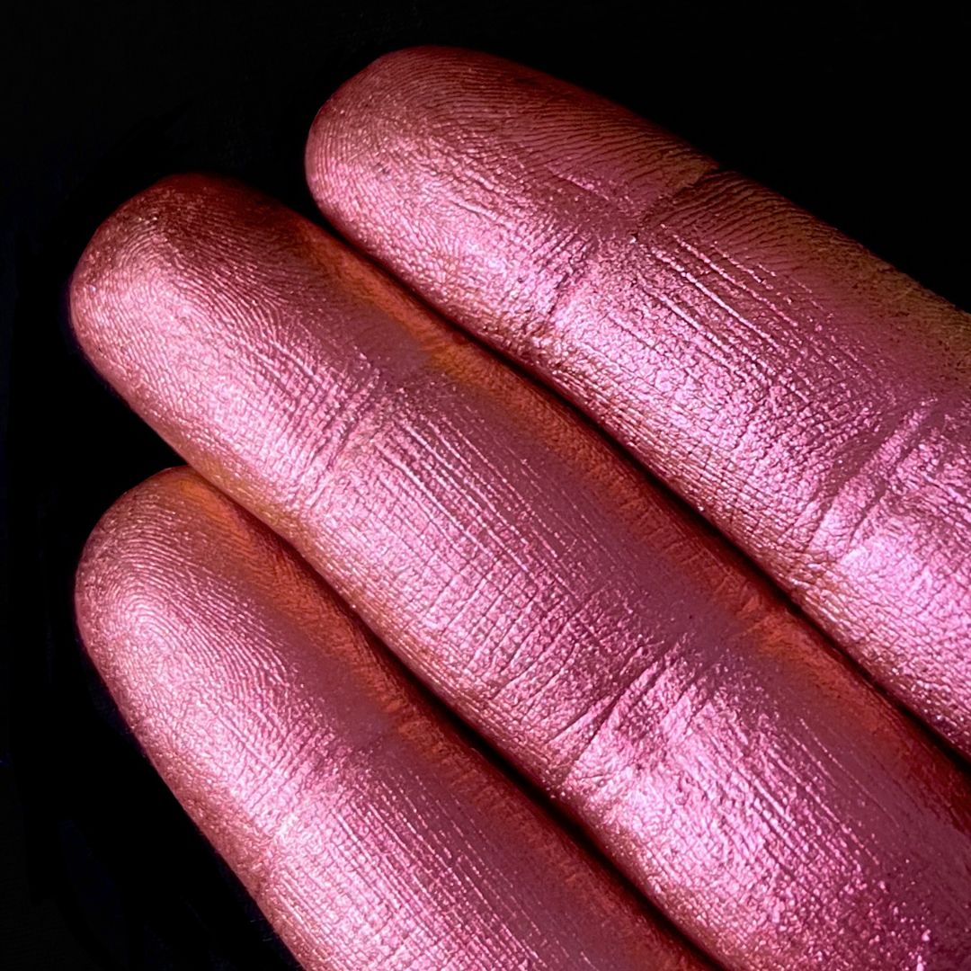 Three fingers display iridescent pastel pink pigment on their nails against a solid black background.