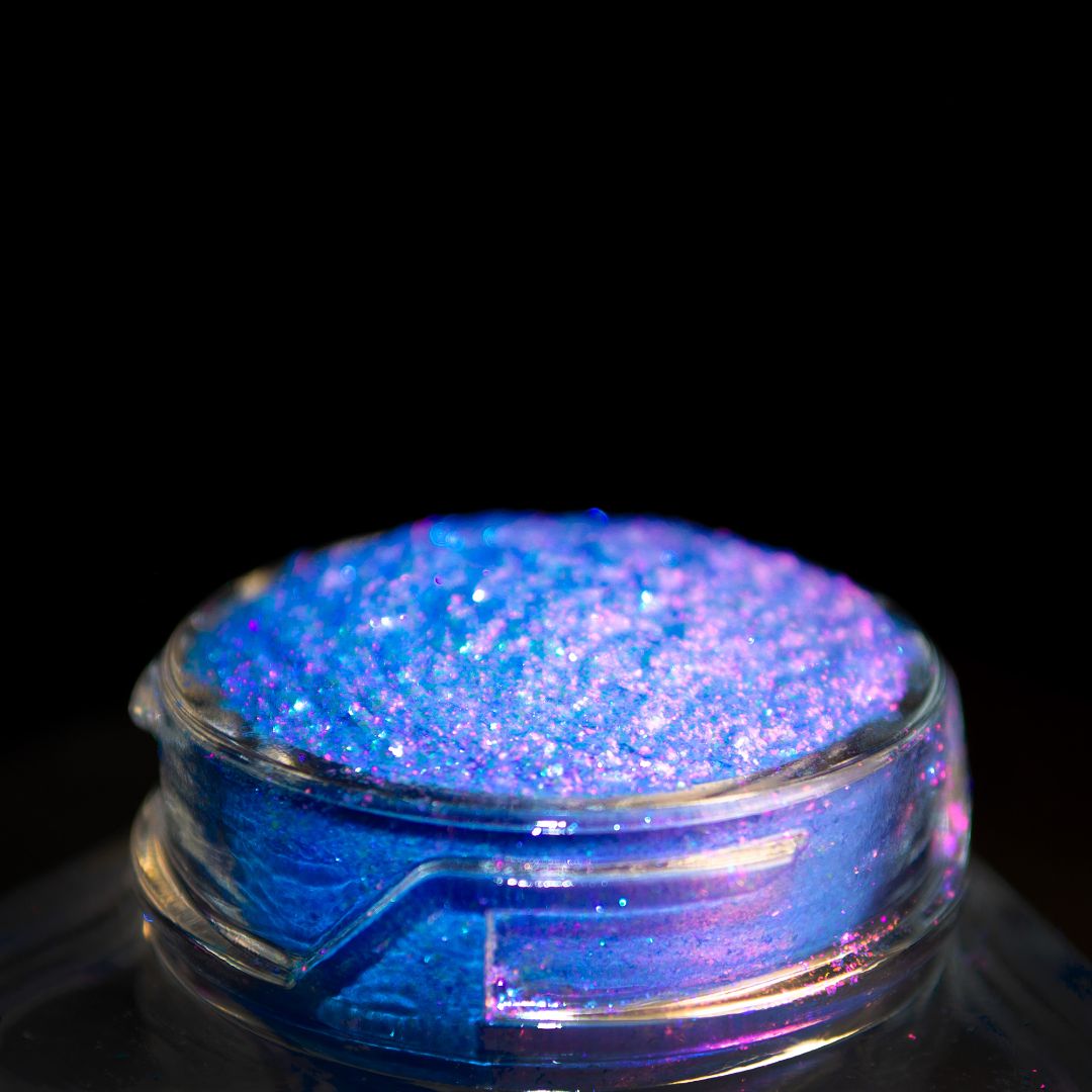 A small glass jar filled with blue and purple shimmer pigment sits against a black background.