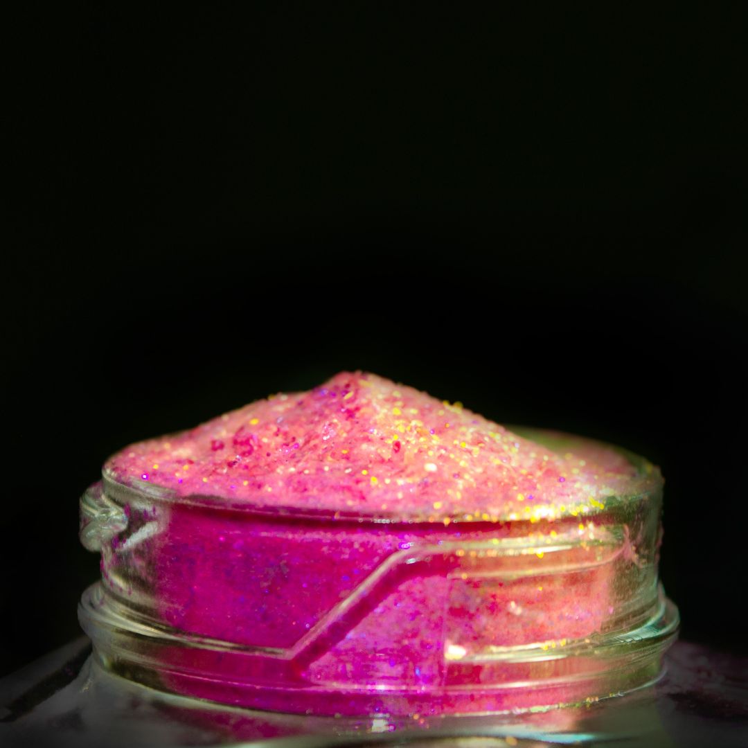 A small glass jar filled with bright pink and gold holographic powder sits on a black background.