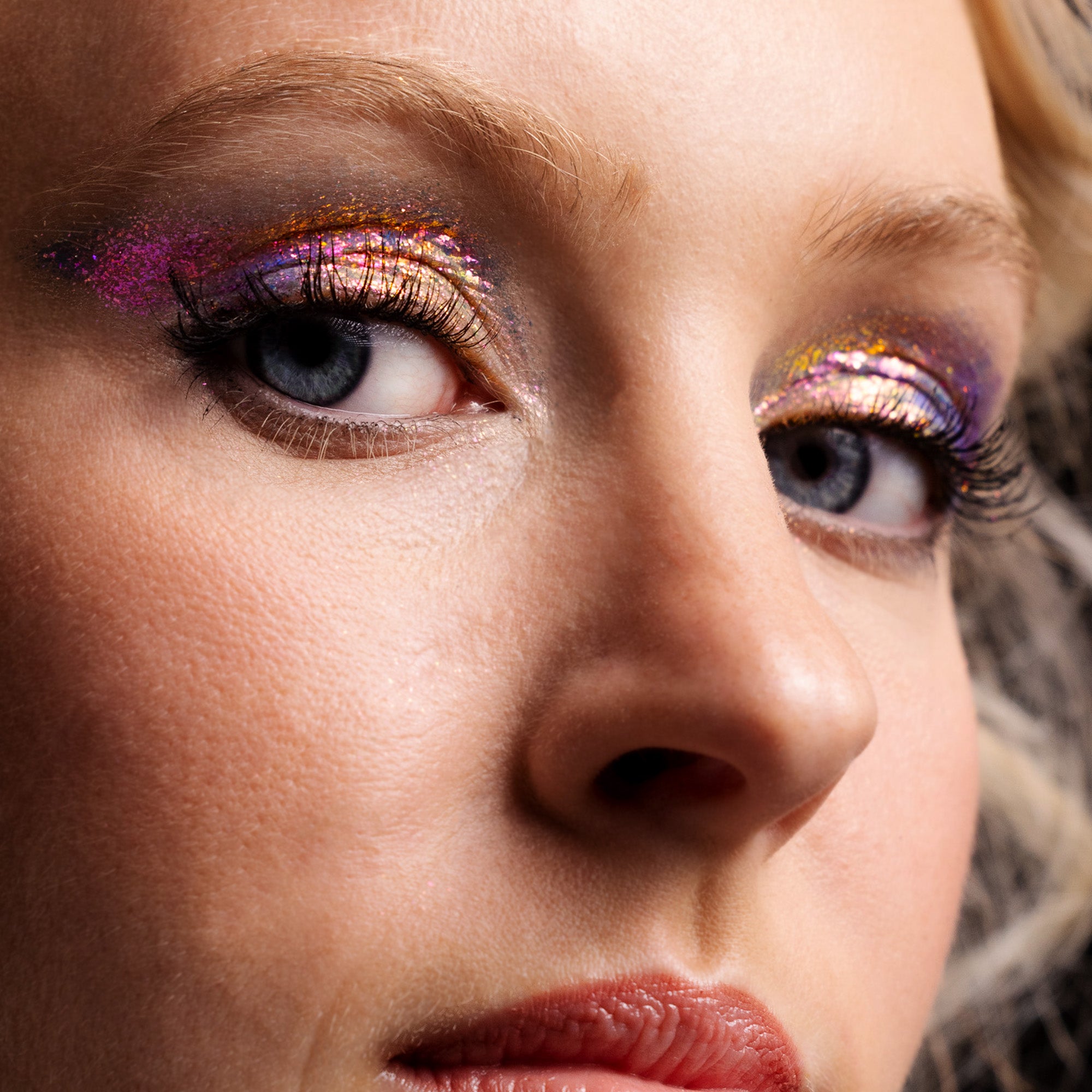 Close-up of a woman's face with glittery eye makeup in purple, gold, and pink shades.