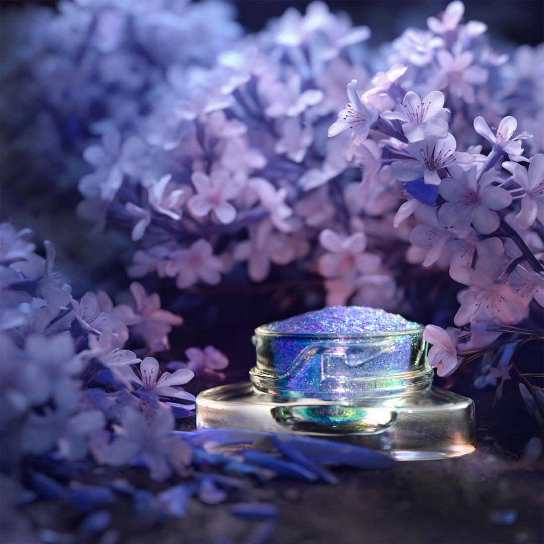 A small glass jar containing iridescent purple glitter is placed among purple flowers in soft lighting.