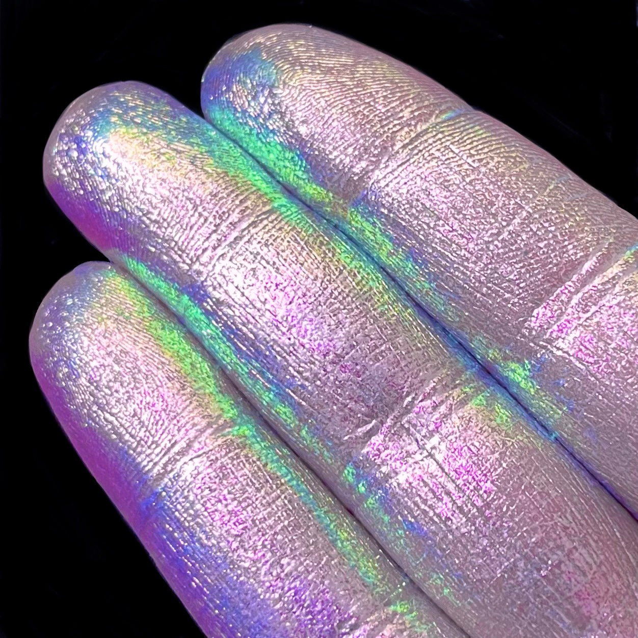 Three fingers display iridescent pastel pigment on their nails against a solid black background.