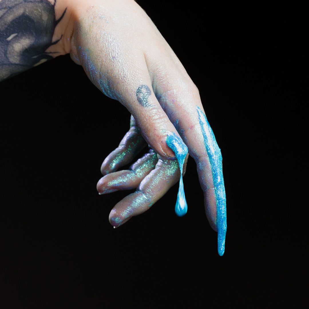 A hand with turquoise glittery paint dripping from fingers against a black background.