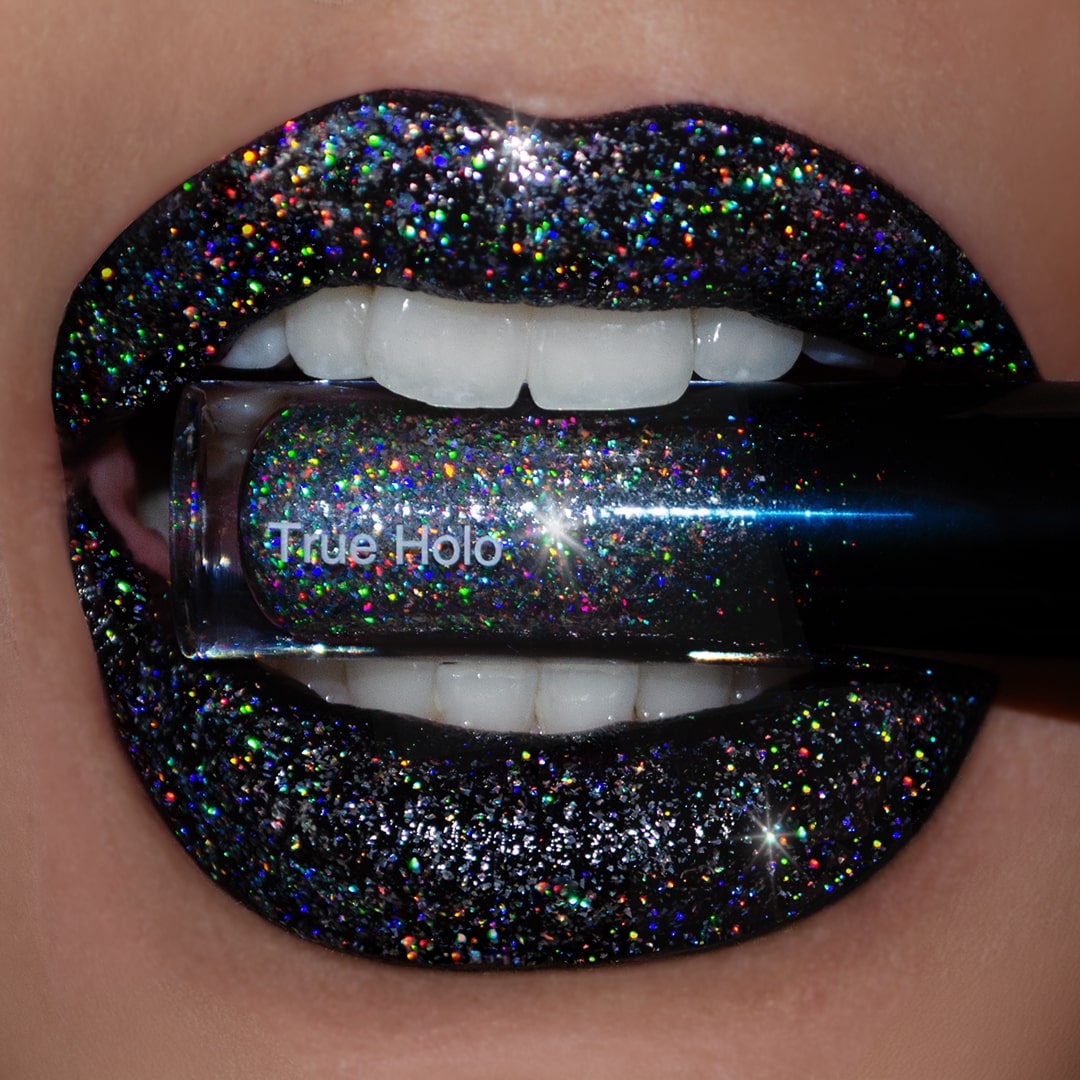 Close-up of lips with black holographic lip gloss and the True Holo tube held between white teeth.