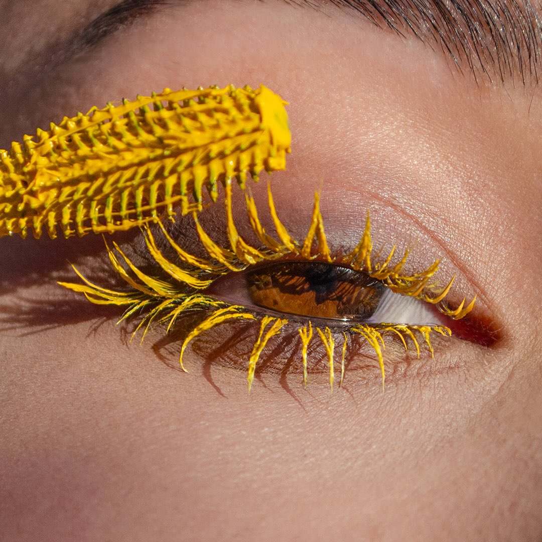 Close-up of an eye with bright yellow mascara on the upper lashes, applied using a spiky wand.