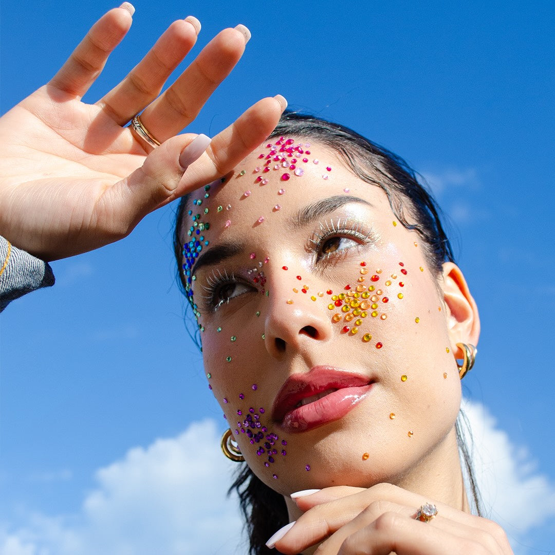 A woman with rhinestones on her face and raised hand stands under a blue sky wearing visible Mascara Arctic Ice.
